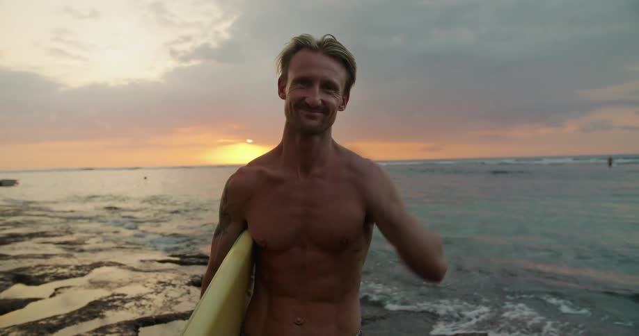 A man is standing on a beach holding his surfboard as waves roll in and the sun sets in the distance