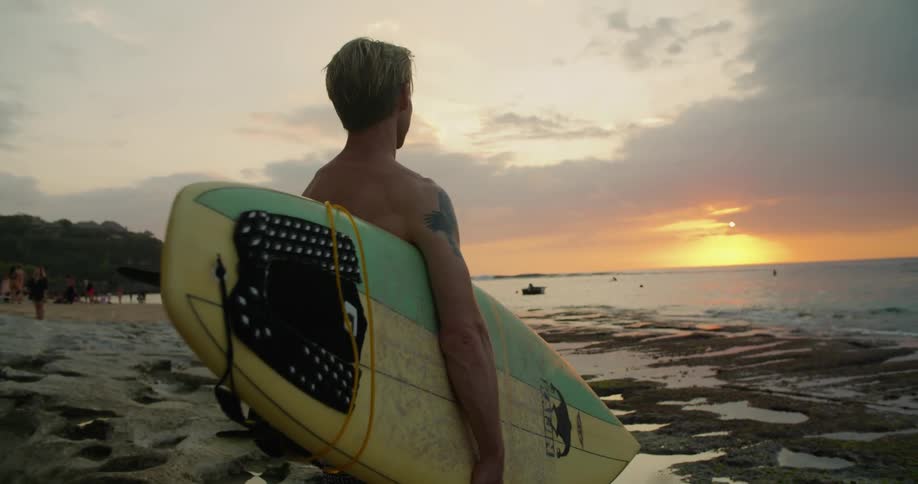 A man standing on a beach with a surfboard at sunset