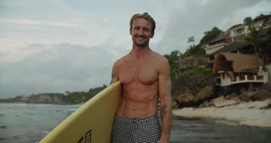 A man is standing on a beach holding his surfboard as waves roll in and there are buildings on the hillside in the distance