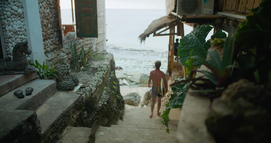 A man walks down the steps out of a building holding his surfboard and he stops to look at the beach and the waves rolling in