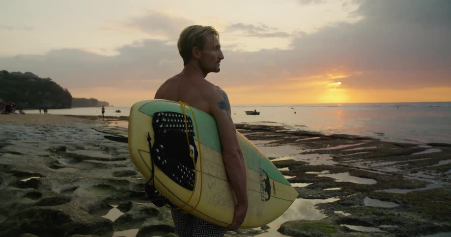 Man holding a surfboard stands on the wet rocks next to the beach at sunset