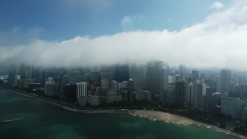 Aerial orbiting view of the skyline of a waterfront city covered in low white clouds at daytime