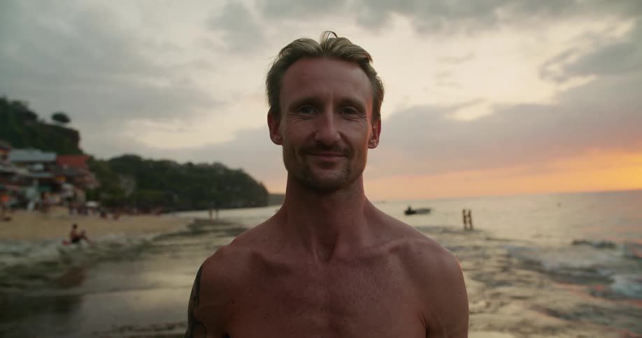 Shirtless man holding a surfboard on a beach at sunset looking at the camera