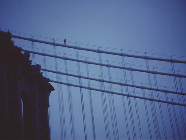 Person is silhouetted against sky while they walk along large supports connected to tower of suspension bridge