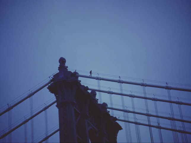 The silhouette of a person walking on the top cables of a bridge against a dark blue sky