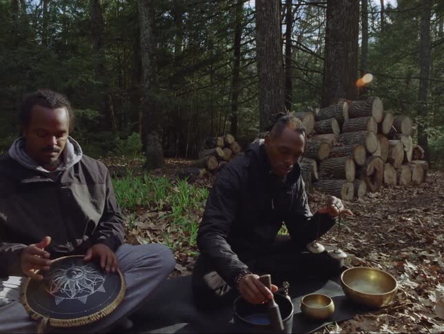 Two men sitting on the ground one playing a lap drum and the other playing singing bowls