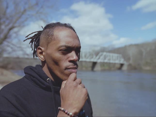 A man chats while standing along a riverbank near a bridge