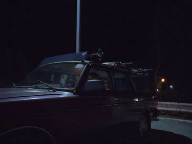 A truck is in a parking lot with the headlights on and the driver leans back in his seat
