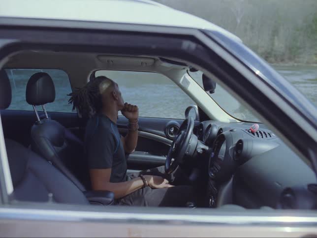 A man gets into an automobile parked along the side of a river and puts on his seat belt