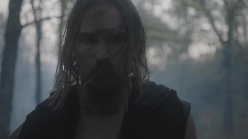 A bearded man stands in a forest