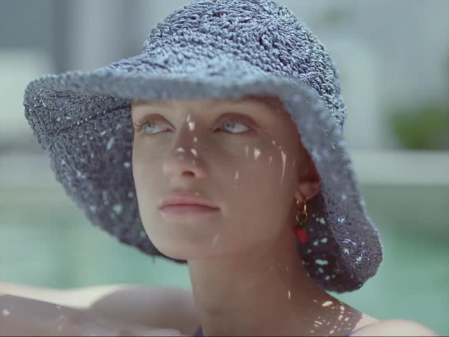 A young woman sitting by the pool wearing a blue hat turns to look at the camera and then looks around