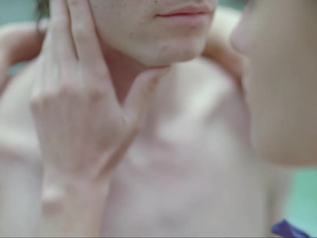 Couple stand facing each other near the water as one touches the others face