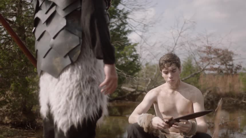 A young man takes a large knife from a larger man while sitting at the edge of a lake