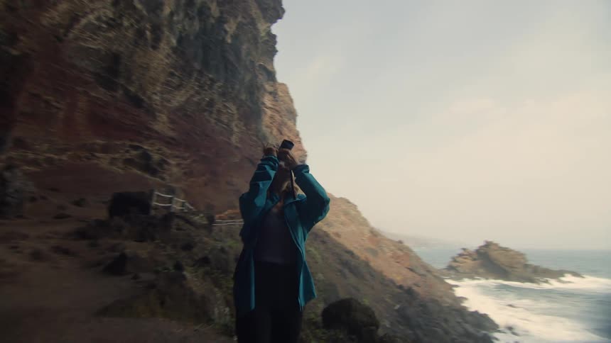 A photographer standing on a path beneath a cliff raises her camera to photograph the cliff above her