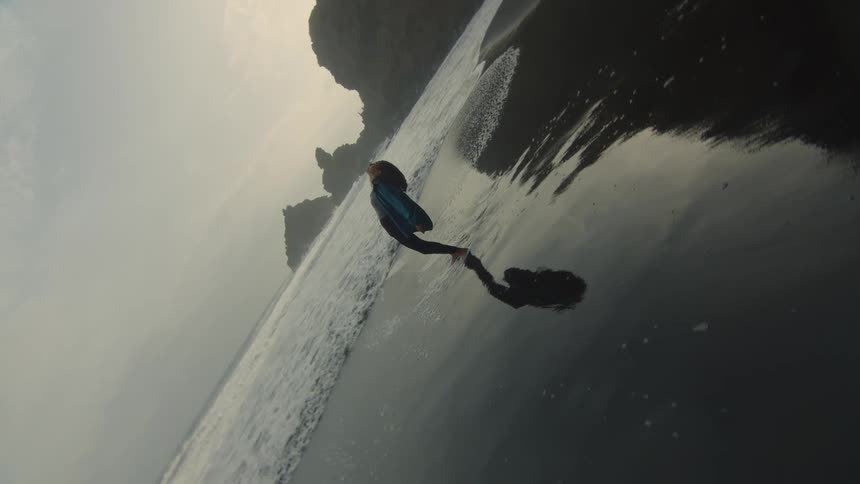A woman is standing on a beach with her feet in the water and she bends her head back looking up and keeps bending backwards