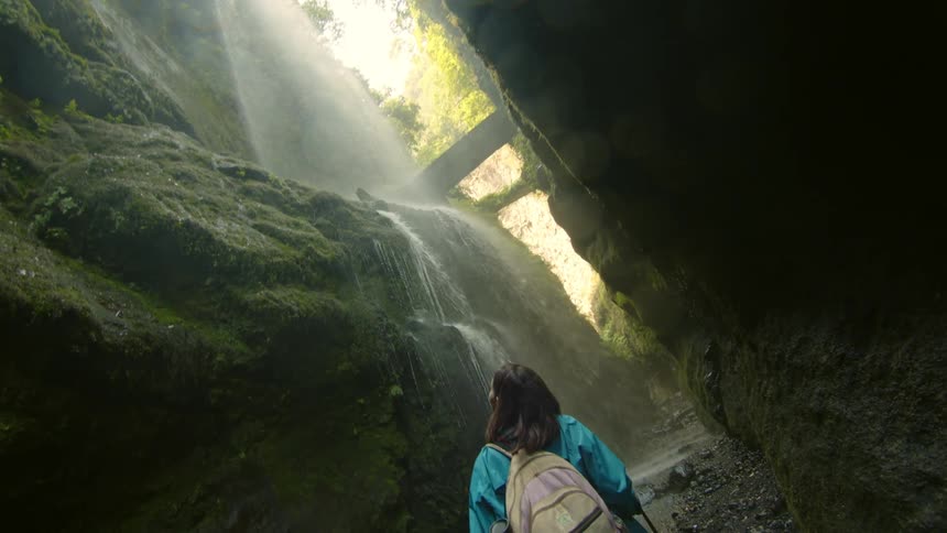 A girl hiking looks up at a waterfall rushing down a rock face
