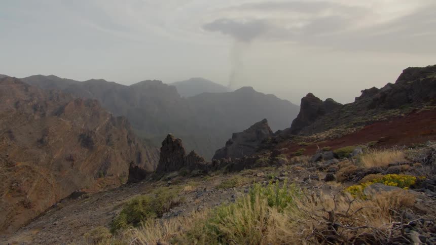 A rocky slope with brush and a plume of smoke rising up into the sky from a mountain ridge