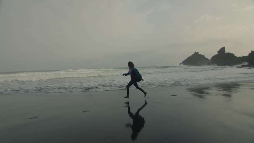 A woman is running barefoot across a beach and leaving footprints in the wet sand as waves roll into shore