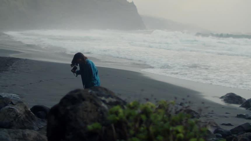 A woman is walking barefoot on a beach carrying her camera and looking around as the waves roll in to shore