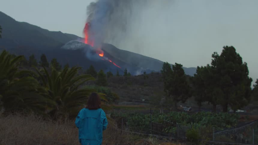 A girl walks forwards as she watches flames and smoke rise from a volcano