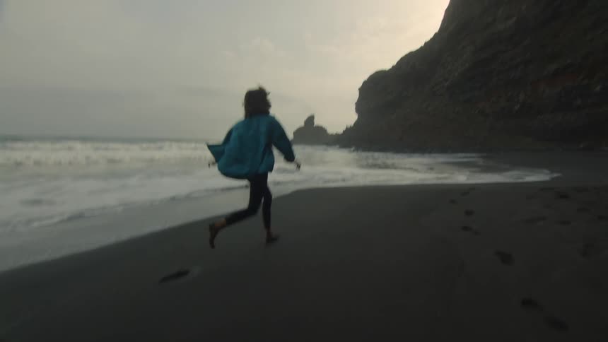 A woman is running barefoot across a beach and leaving footprints in the wet sand as waves roll into shore