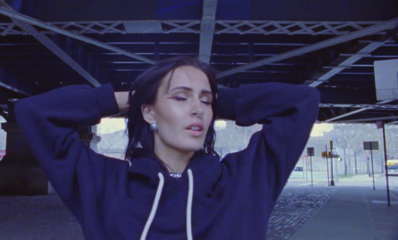 A woman wearing a hoodie jogs a bit under the elevated train tracks then stands with hands on her head looking around