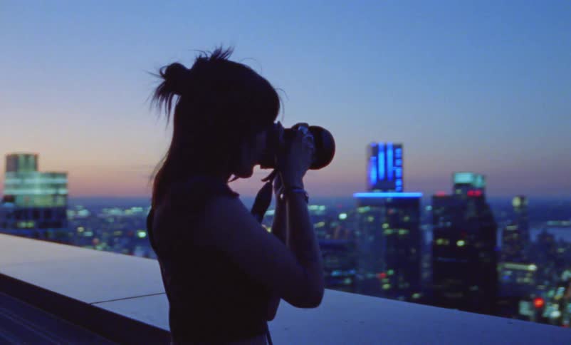 A woman is standing on a balcony taking pictures of the city with lights on the buildings