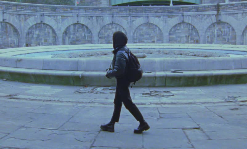 A woman walks down a sidewalk and stops by an empty fountain and takes a picture