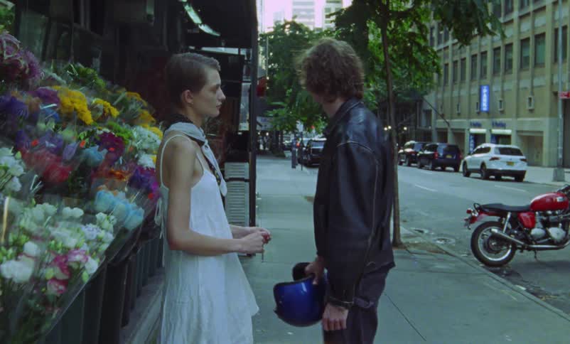 A man is standing on a sidewalk holding a womans hands as she stands in front of a flower display she leans over and kisses him and he walks to his motorcycle parked in the street