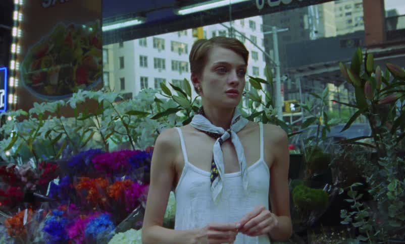 A woman standing in front of a flower shop in a city raises her hand up and rests her face in it