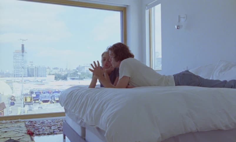 Man with tattoo white shirt curly hair and gray pants lies on bed with woman touching hands in bedroom