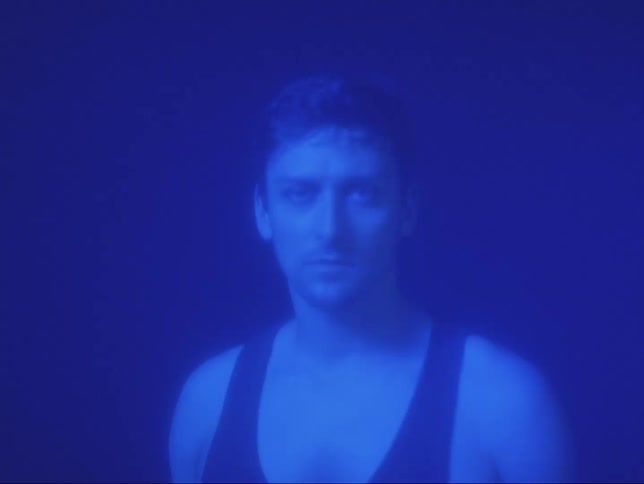 A man bathed in a blue light stands staring