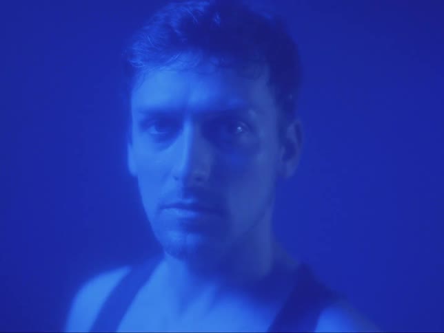 A man turns around but stares straight ahead in a dim room with a blue light