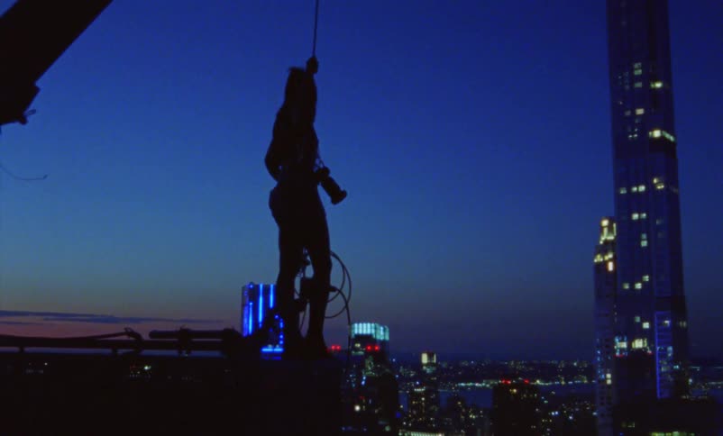 The silhouette of a woman holding a construction crane rope with her camera around her neck high above the city
