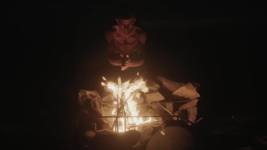 Shirtless man warming his hands over a campfire at night