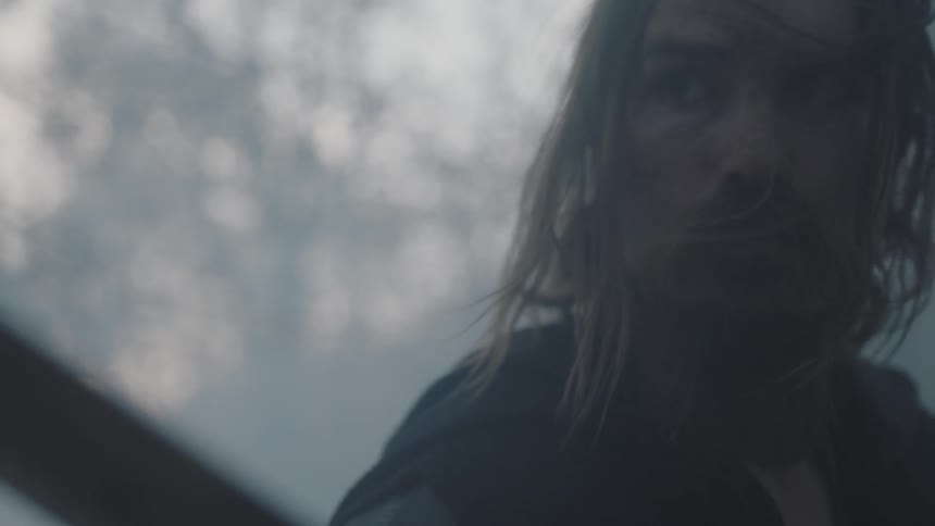 A man with long hair raises a piece of wood towards an unknown subject