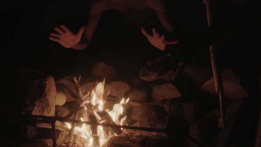 A person stirs a fire and warms their hands at a campfire