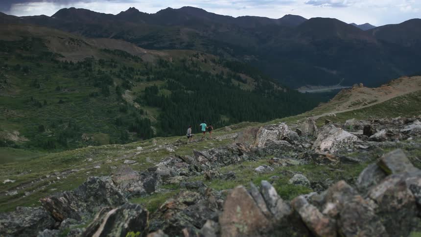 Three people are running down a path on a rock covered hillside and there are mountains in the distance