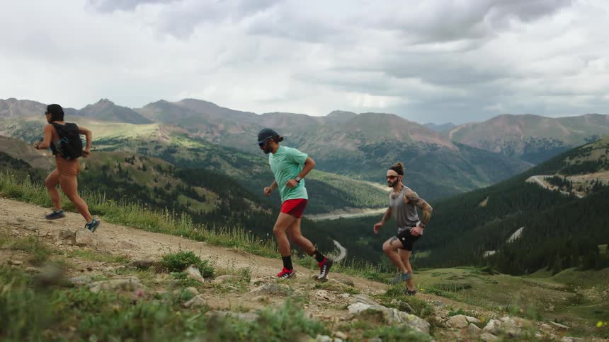 Three people jogging and running up a sloped trail with mountain ridges and cloudy skies on the horizon