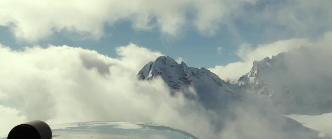 Mountains with peaks high in the clouds are seen through an airplane window as the propeller spins
