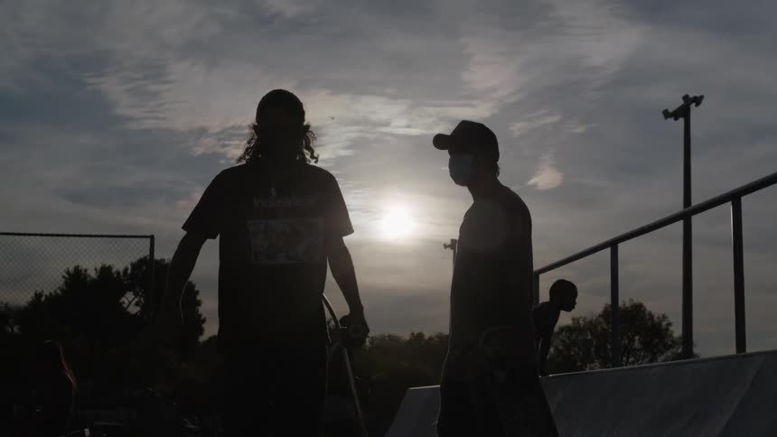 Two friends fist bump at a skateboard park as young kids climb up the wall behind them