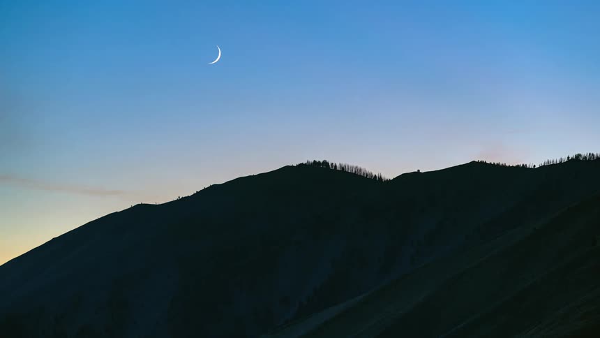 Crescent moon sits over red tinted sky and nearly silhouetted slope
