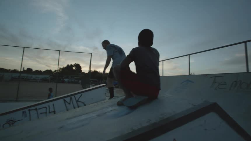 A teenager boy is standing on top of a ramp at a skateboard park and he pushes his skateboard down and glides down the pavement as another kid watches
