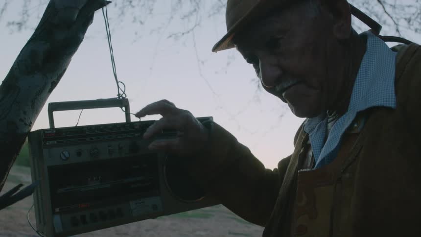 An old man bends his head to listen as he tunes a radio hanging from a tree branch