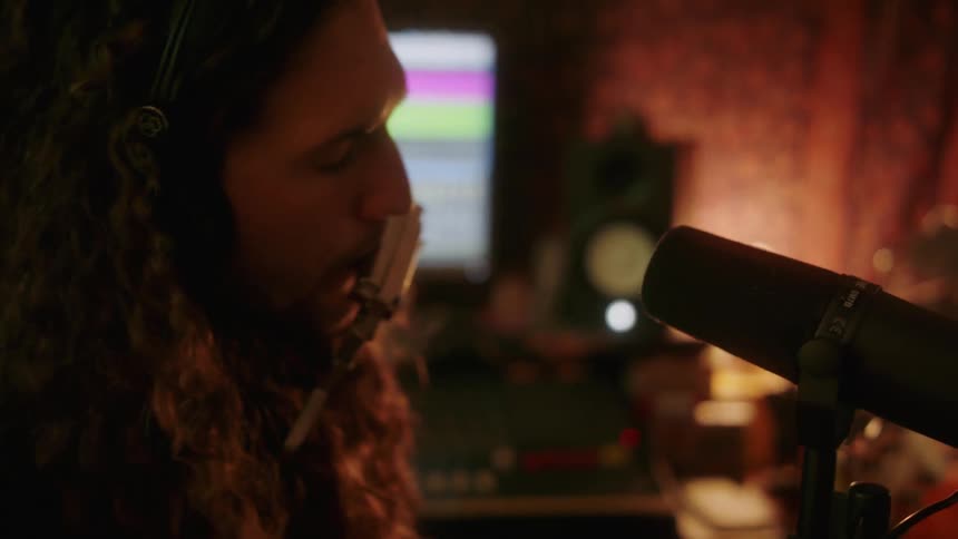 A man is sitting down in a studio playing a guitar and singing