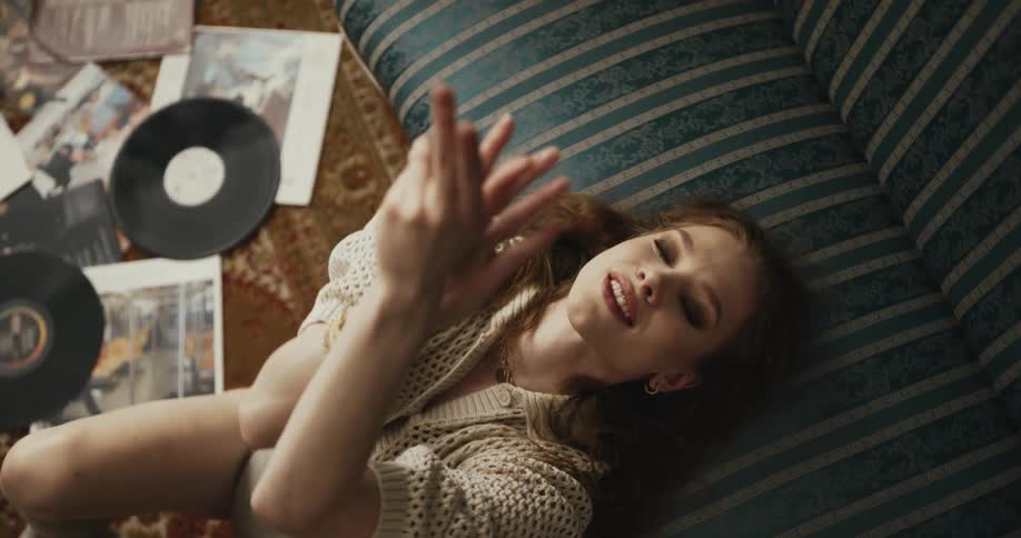 Woman leans with her head on sofa seat and gestures with her hands as she sits on floor near vinyl records