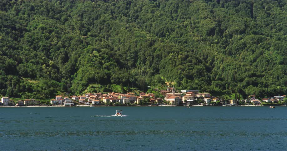 A boat travels along the coast of a small town at the base of a forested mountain