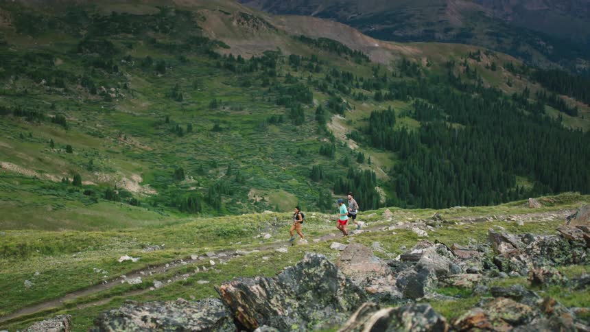 Runners run along dirt trail through valley with forest dappled mountains in the background