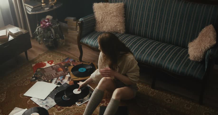 Woman with brown hair necklace holds lp record sitting in front of blue couch smiling and dancing