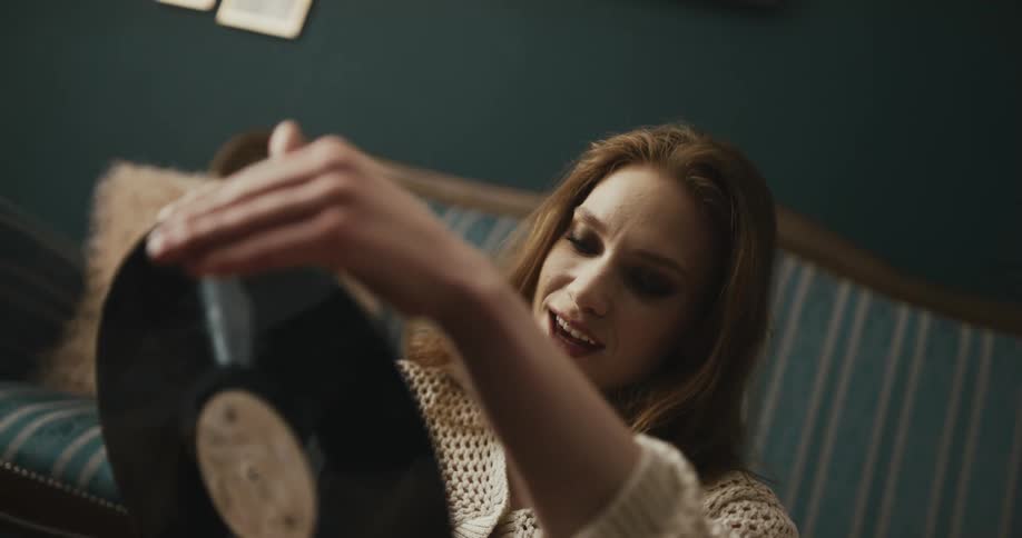 Woman with brown hair necklace holds lp record sitting in front of blue couch smiling